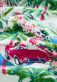 Czerwony samochód kabriolet jadący przez tropikalną scenerię z palmami, niebieską rzeką i kolorowymi budynkami na tkaninie.