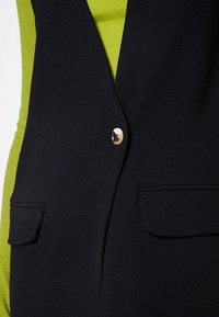 Svart väst med en enda guldknytnad, två framfickor och en mjuk textur. I kombination med en ribbad neongrön topp under.