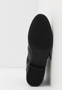 Chaussure en cuir noir avec une semelle texturée et des accents cloutés. La chaussure est inclinée pour mettre en valeur le dessous plat et lisse ainsi que les détails.
