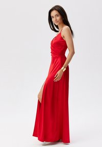 Dlouhá červená saténová šaty s upnutým živůtkem, tenkými ramínky a bočním rozparkem. Vyznačují se hladkou texturou a elegantním splýváním. Doplňují je zlatý náramek.