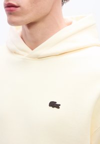 Άτομο που φοράει φούτερ με κουκούλα κρεμ χρώματος, με μικρό σκούρο κεντητό λογότυπο κροκόδειλου στο στήθος, εμφανίζοντας μερικώς τον λαιμό και τα χείλη.