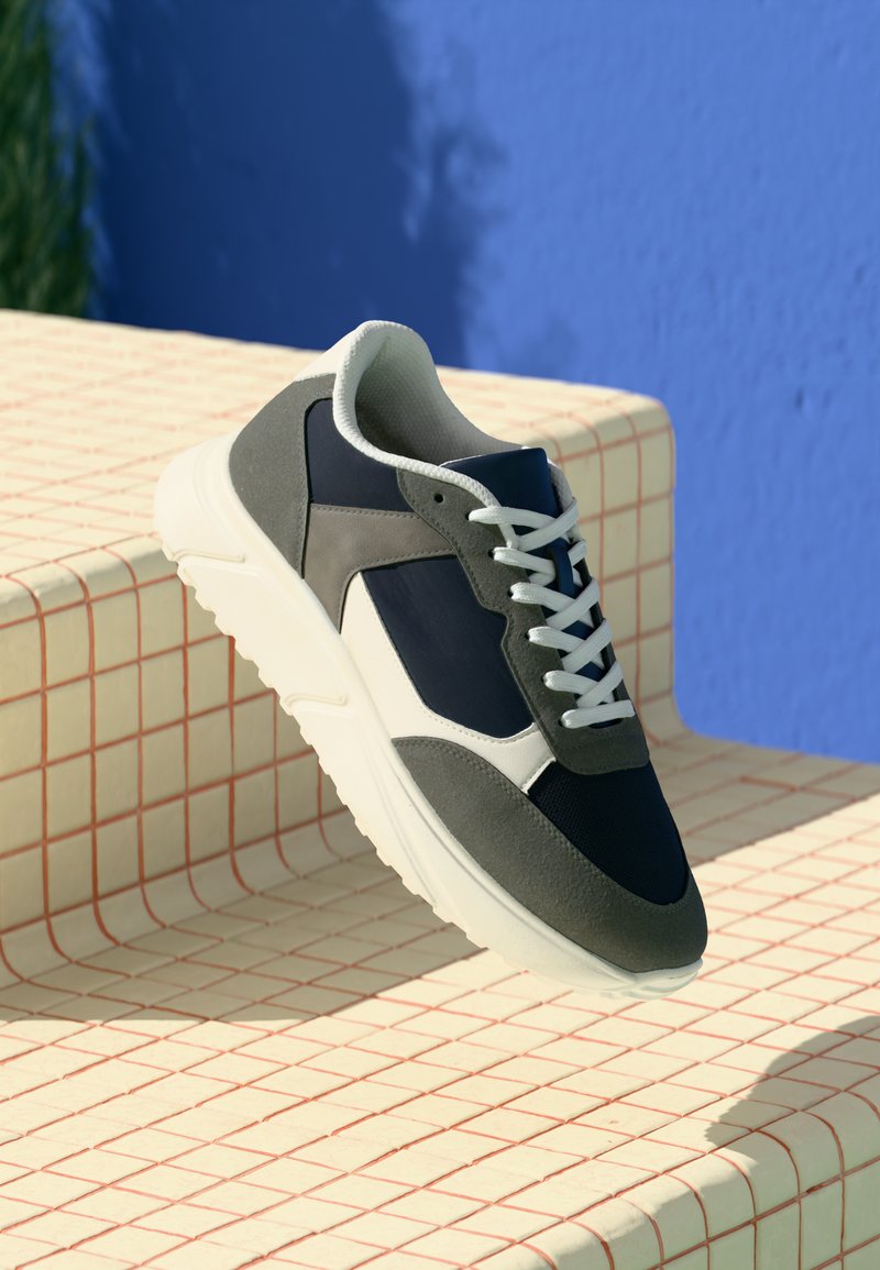 Gray and navy sneaker with white laces, featuring textured fabric and smooth accents. Set on a patterned platform against a blue background.