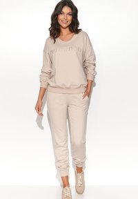 Beżowy zestaw bluzy i spodni dresowych, luźny krój z prążkowanymi mankietami, z subtelnym, lśniącym napisem "numinou". Miękka tekstura materiału.