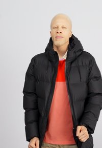 Zwarte pufferjack met een ritssluiting aan de voorkant, grote capuchon en elastische manchetten, gedragen over een shirt in rood en roze blokken.