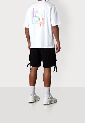 Homme portant un t-shirt blanc oversize avec un texte graphique coloré, un short cargo noir, des chaussettes blanches et des baskets blanches, debout de dos.
