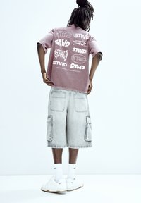 T-shirt oversize rosa con vari loghi sulla schiena, abbinata a pantaloni cargo grigi e sneakers bianche. Calzini bianchi visibili.