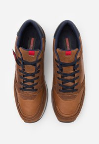 Brown leather sneakers with navy accents, featuring a lace-up design, stitched detailing, and a textured surface. Red tab on side.