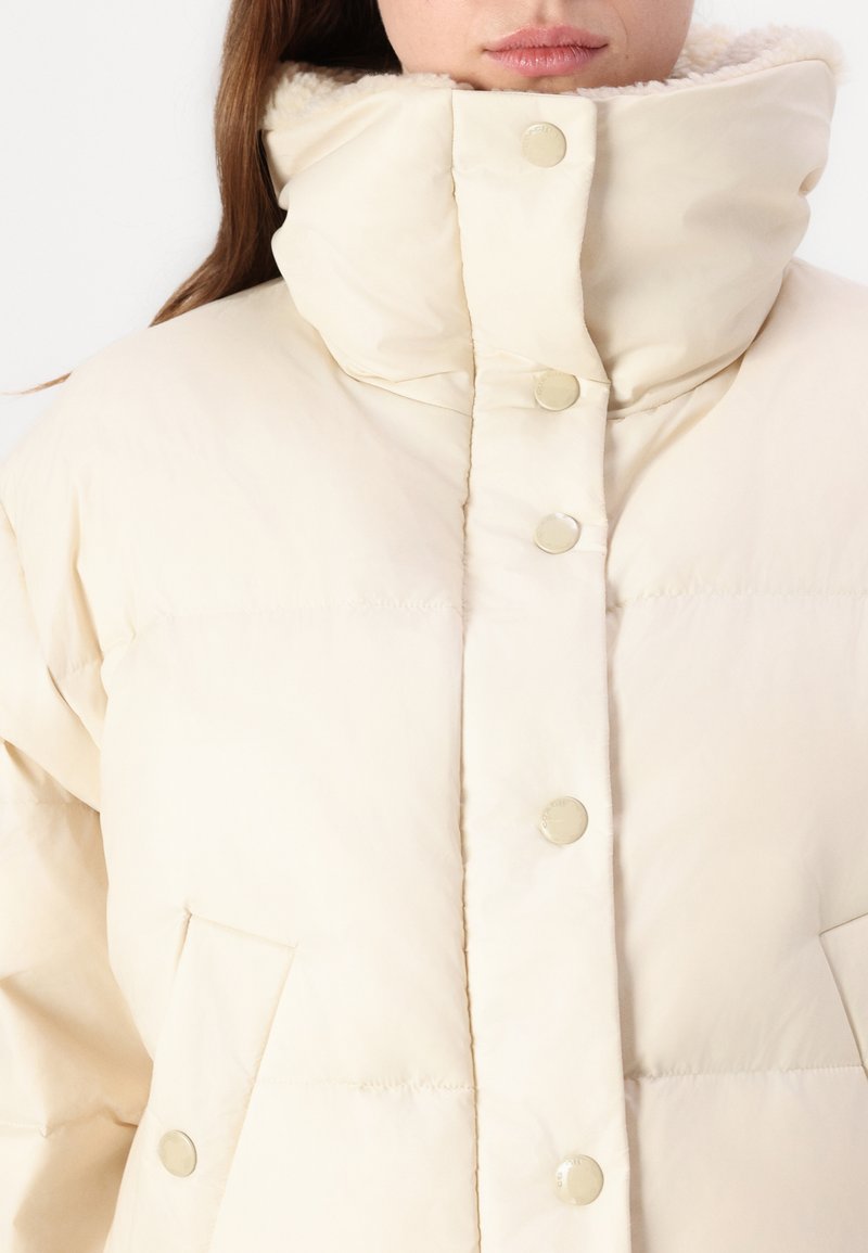 Cremfarbener, gesteppten Pufferjacke mit hohem Kragen, Druckknöpfen und weichem Fleece-Futter. Verfügt über seitliche Taschen und eine lockere Passform.