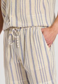 Lätt, stickad koordsett i krämfärg med blå vertikala ränder. Innehåller knappar på toppen, elastisk midja och sidofickor.