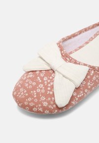 Chausson en tissu rose avec un petit imprimé floral blanc et un grand nœud en tissu blanc sur le bout, doublure intérieure douce visible.