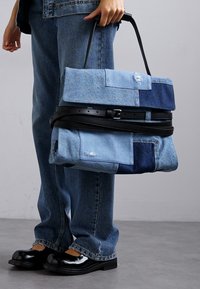 Torba od trapera s patchwork dizajnom u svijetloplavim i tamnoplavim nijansama, zatvorena crnim remenom. Nošena uz odgovarajuće traperice.