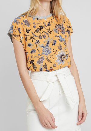 Woman wearing a yellow floral patterned short-sleeve shirt tucked into a high-waisted white skirt with a belt tied at the waist.