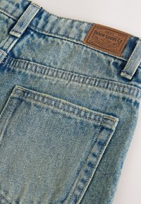 Jean en denim bleu clair avec une finition texturée, comportant une étiquette en cuir portant la mention "DENIM GOODS CO" et des détails de poche arrière cousus.