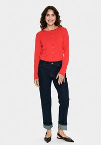 Raudona ilga rankovė kardiganas su mygtukais, derinamas su tamsiai mėlynais įmantrių rankovių džinsais ir rudomis plokščiomis bateliais. Lygus audinys, laisvas siluetas, minimalus detalizavimas.