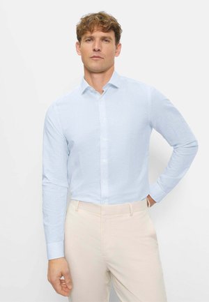 Mann trägt hellblau gestreiftes, langärmliges Hemd mit Knöpfen und beige Hose, steht mit einer Hand in der Hüfte vor weißem Hintergrund.