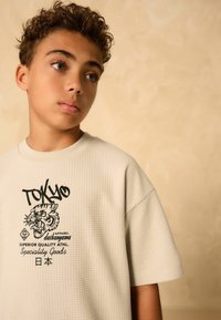 T-shirt a maniche corte color beige chiaro con un motivo a griglia testurizzato, caratterizzato da una grafica di una tigre nera e il testo "TOKYO" sul davanti.