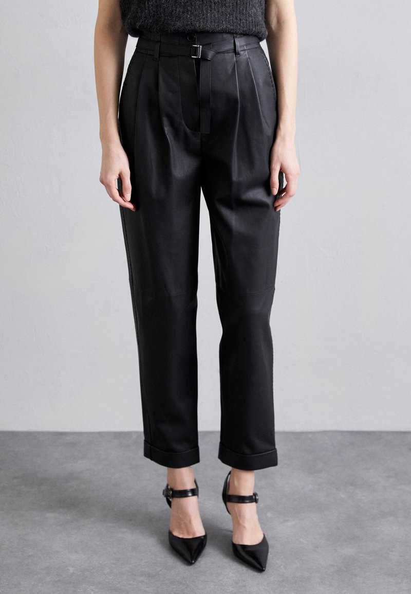 Μαύρο δερμάτινο παντελόνι ψηλής μέσης με πιέτες, διαθέτει ζώνη με αγκράφα και γυρισμένα μπατζάκια, συνδυασμένο με μαύρες μυτερές γόβες.