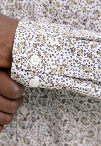 Camicia bianca con motivo floreale in tonalità beige e marrone, con polsini arrotondati e chiusura a bottoni bianchi. La texture è liscia e leggera.