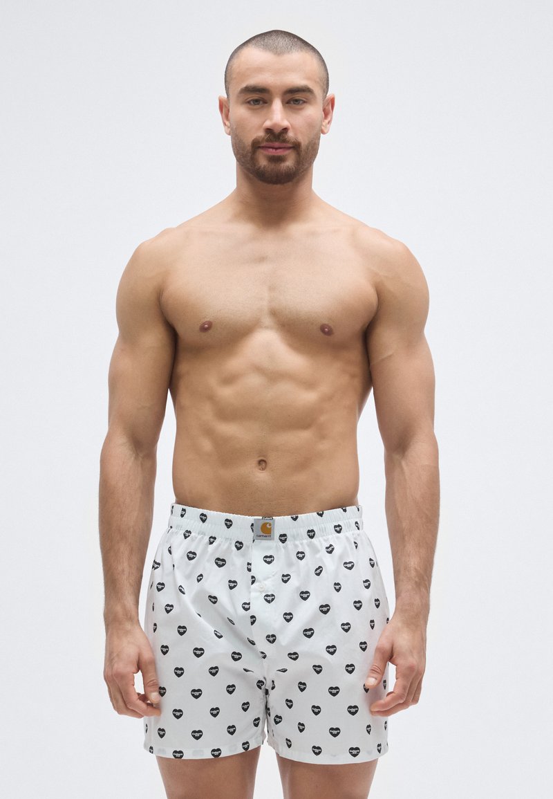 Boxer bianchi con motivi a cuori neri, vita elastica con etichetta logo. Modello in piedi senza maglietta, rivolto in avanti. Texture liscia.