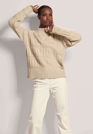 Pull en tricot beige avec des poignets et un ourlet côtelés, présentant un motif en losange texturé. Associé à un pantalon en velours côtelé blanc.