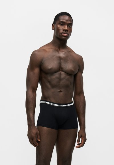 Mannequin masculin noir musclé debout portant un boxer noir Hugo avec une ceinture blanche sur un fond gris clair uni.