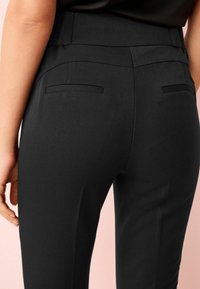 Pantalon noir avec une coupe ajustée, présentant une texture lisse, des poches arrière élégantes et une taille mi-haute. Pas de logos ou motifs visibles.
