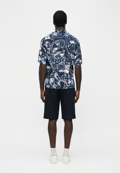 Άνδρας φοράει πουκάμισο με κοντά μανίκια σε ναυτικό μπλε και λευκό με μοτίβο λουλουδιών, μαύρα σορτς, λευκές κάλτσες και λευκά αθλητικά παπούτσια, στέκεται με την πλάτη στραμμένη.