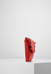 Rød læder clutchtaske med en bølgende flap. Designet er slankt med en lynlås-lukning og en glat overflade, stående oprejst på en hvid overflade.