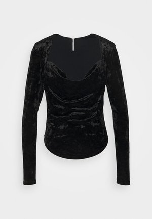 Top de terciopelo negro de manga larga con escote redondeado, que presenta detalles fruncidos y un dobladillo ligeramente curvado. Cierre de cremallera en la parte trasera.