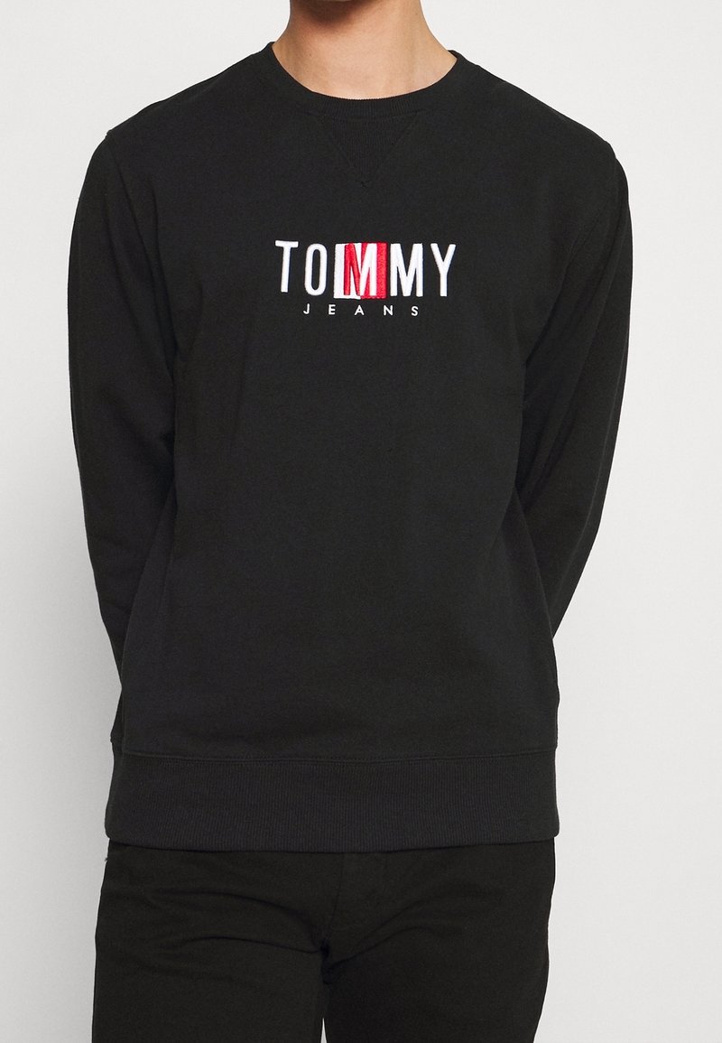 Personne portant un sweat-shirt noir à manches longues avec le logo "TOMMY JEANS" en blanc et rouge centré sur la poitrine.