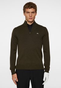 Tummanoliivinvihreä vetoketjullinen neule, jossa on ribbausta hihansuissa ja helmassa. Rintapielessä pieni valkoinen logo. Päällä musta paita.