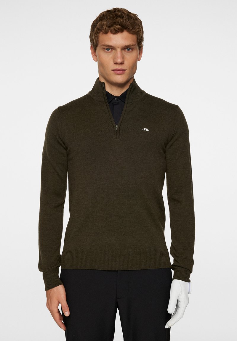 Tummanoliivinvihreä vetoketjullinen neule, jossa on ribbausta hihansuissa ja helmassa. Rintapielessä pieni valkoinen logo. Päällä musta paita.