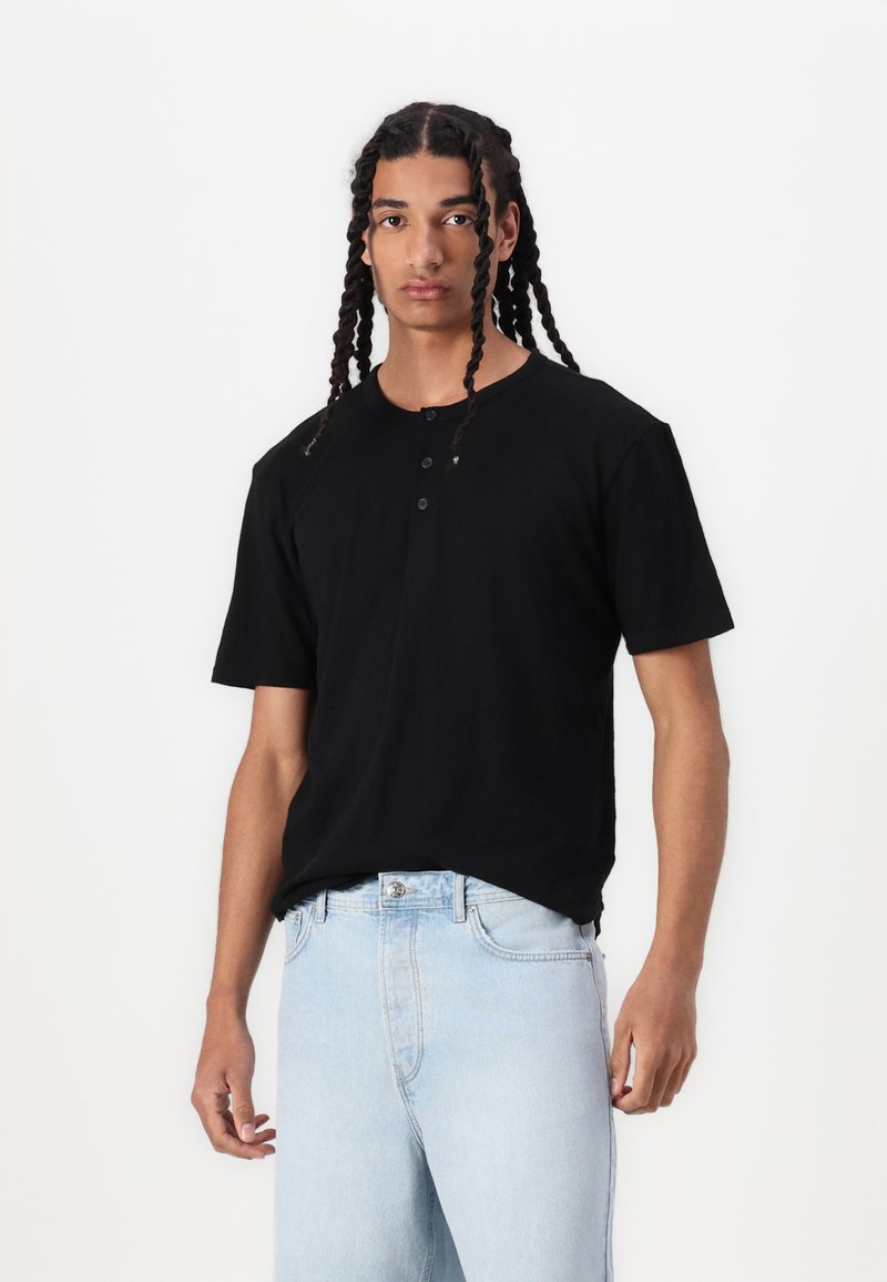 Schwarzes Baumwoll-T-Shirt mit kurzen Ärmeln und Drei-Knopf-Leiste, lockere Passform, kombiniert mit hellblauen Jeans. Glatte Textur, simples Design.