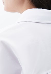 Camicia bianca con un tessuto morbido e leggero. Il primo piano si concentra sul colletto e sulla spalla, mostrando una texture minimale e linee pulite.