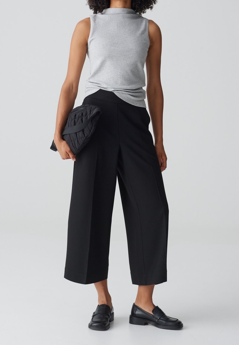 Haut gris sans manches et pantalon large noir. Le pantalon a une texture lisse, tandis que le haut présente un drapé subtil. Un pochette noire matelassée est tenue en main.