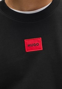 Svart sweatshirt med rund hals, med en röd rektangulär patch med ordet "HUGO" i svarta bokstäver, som visar en strukturerad tyg.