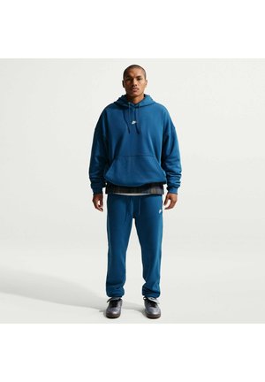 Person står med fronten frem iført en blå Nike-hættetrøje og matchende joggingbukser med sorte sneakers mod en lys ensfarvet baggrund.