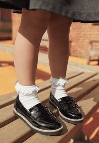 Black patent loafers with textured accents, paired with white ankle socks featuring lace trim, positioned on a wooden surface.