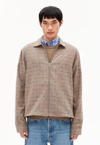 Jongen in een beige geblokte zip-up jack over een bruine trui en een blauw gestreepte blouse, gecombineerd met blauwe jeans, op een witte achtergrond.