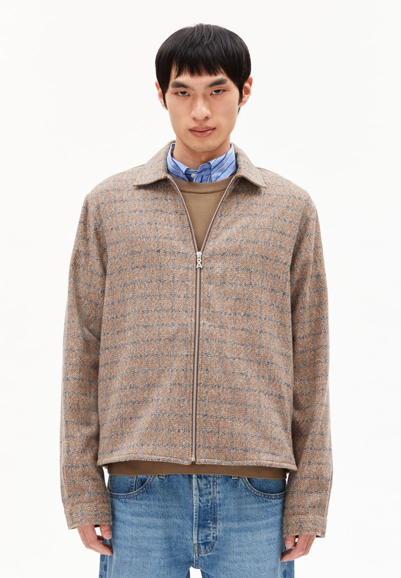 Jongen in een beige geblokte zip-up jack over een bruine trui en een blauw gestreepte blouse, gecombineerd met blauwe jeans, op een witte achtergrond.