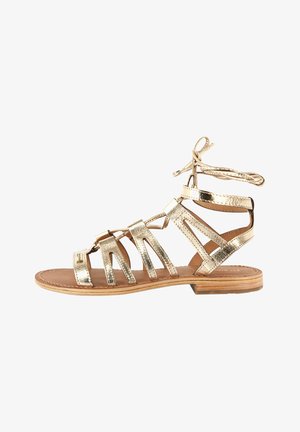 Gouden metallic gladiator sandaal met meerdere banden, enkelbandjes en een platte zool. Bruine leren voetbed met een gladde afwerking.