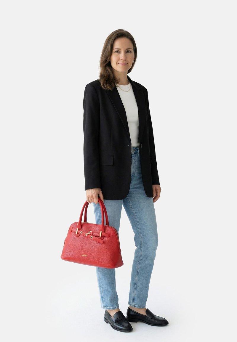 Mujer de pie con blazer negro, blusa blanca, vaqueros azul claro, mocasines negros, sosteniendo un bolso de cuero rojo por las asas.
