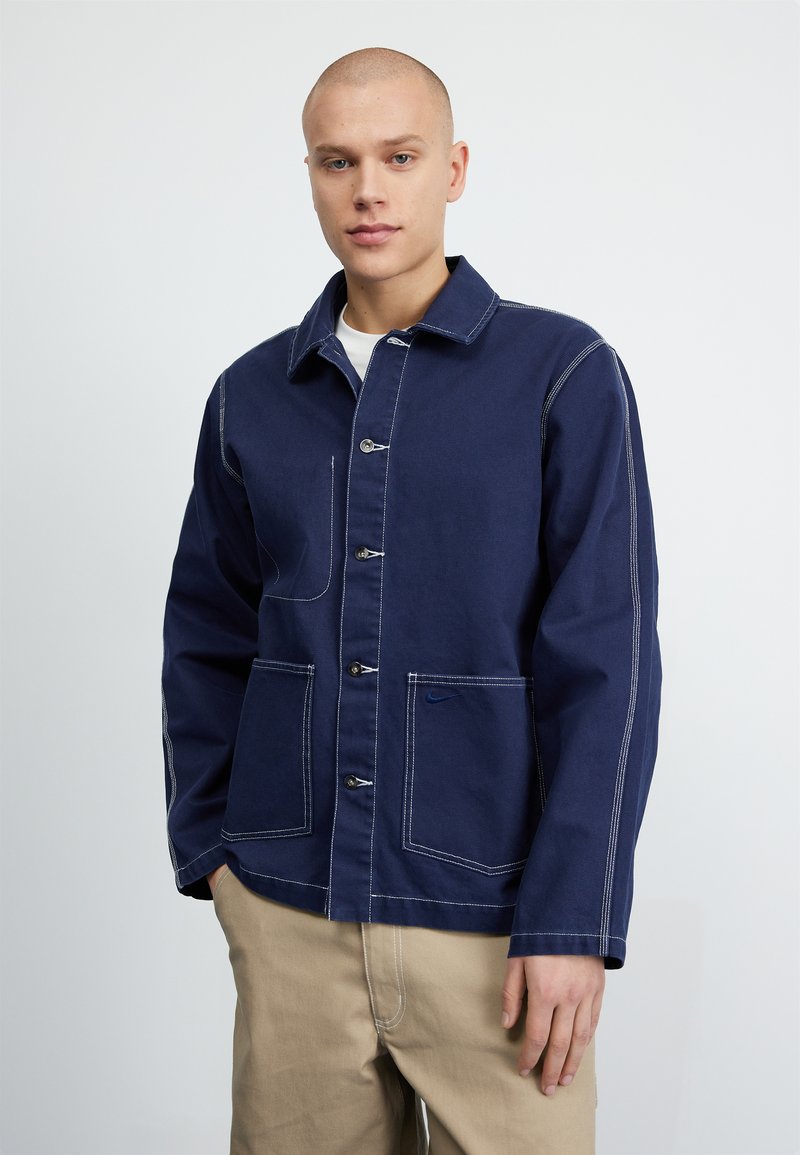 Chaqueta azul marino, material de algodón, diseño abotonado, dos bolsillos delanteros, costuras blancas en contraste y un pequeño logo en el bolsillo.