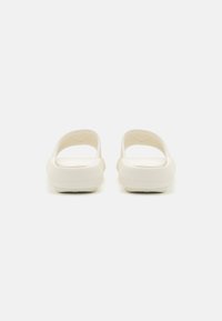 Hvite slip-ons med teksturert materiale, åpen bak, og flate såler. Innvendig har de et ribbet mønster for komfort.