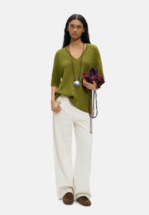 Kobieta ubrana w luźny, oliwkowozielony dziergany sweter, białe szerokie spodnie, brązowe mokasyny, długi naszyjnik i trzymająca fioletową torebkę z frędzlami.