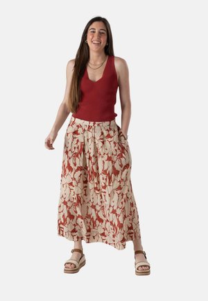 Joven sonriente con camiseta roja sin mangas, pantalones anchos con estampado y sandalias de plataforma beige, posando frente a un fondo blanco.