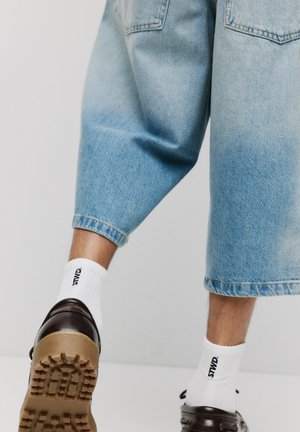 Hellblaue Jeans-Culotte mit weitem Bein und zwei Gesäßtaschen. Getragen mit weißen Socken, die mit "STND" gekennzeichnet sind, und dunkelbraunen Schuhen.