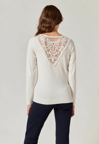 Jasnobeżowy sweter z długimi rękawami; na plecach ażurowy wzór w kształcie litery V. Miękka faktura, ściągacz na dole i przy mankietach.