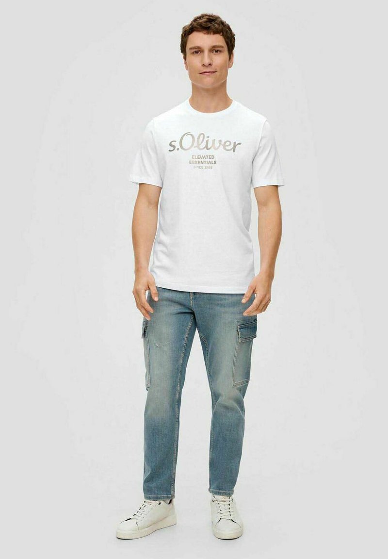 White cotton t-shirt with short sleeves featuring gray text "s.Oliver Elevated Essentials Since 1969." Paired with light blue cargo jeans and white sneakers.