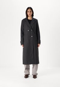 Grijze wollen lange jas met een dubbellaags ontwerp, grote revers en zijzakken, gecombineerd met lichtgrijze broek en donkere schoenen.